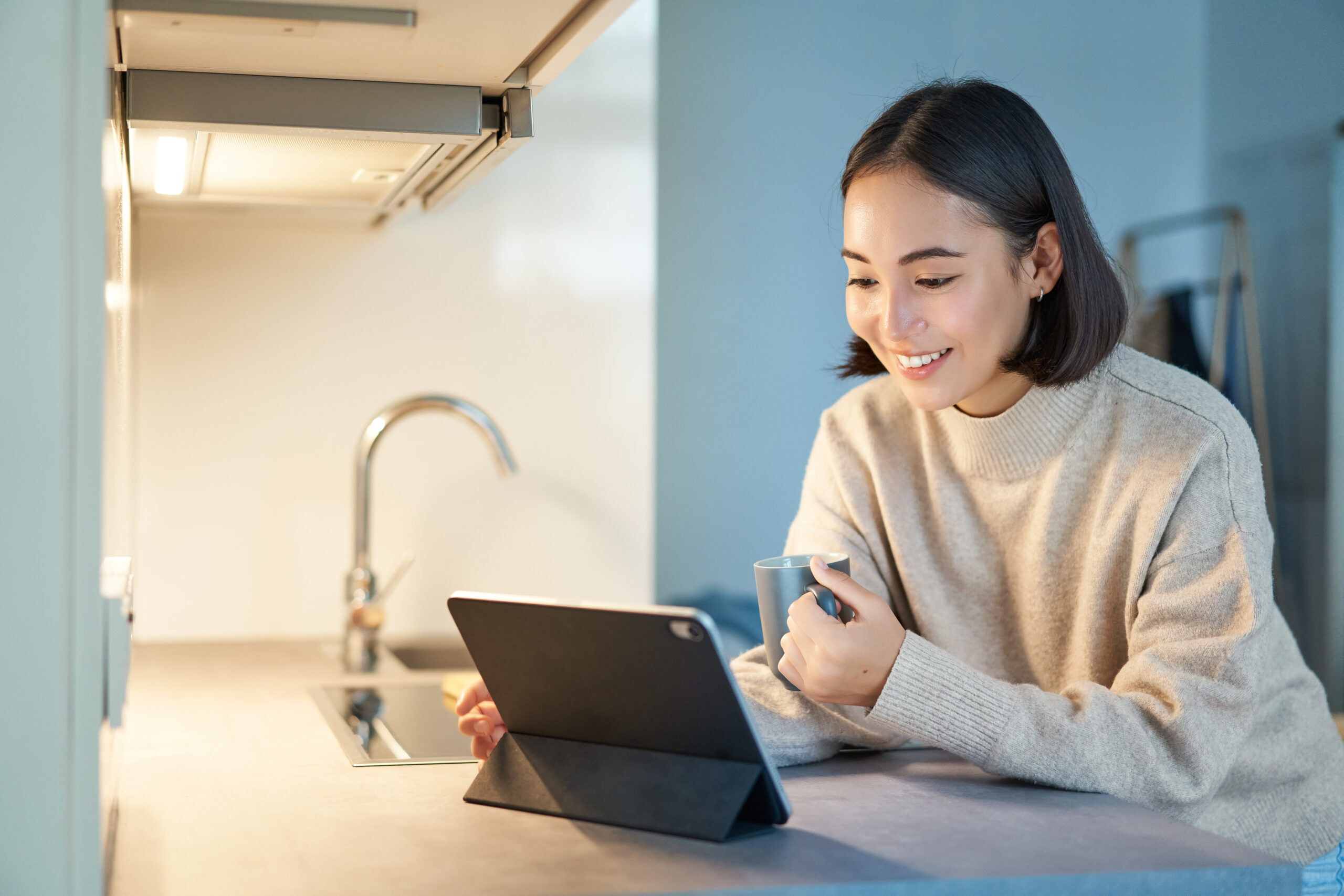 Portrait of stylish young asian woman watching videos on tablet, sitting in kitchen and drinking coffee